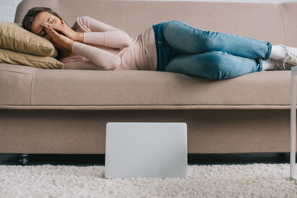 portátil en la alfombra y la mujer joven que sufre de dolor de cabeza en el sofá en casa
 - Foto, Imagen