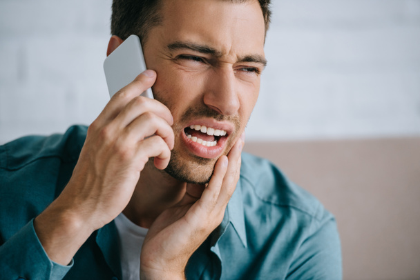 jonge man praten door smartphone en weg te kijken terwijl het lijden aan tand pijn thuis   - Foto, afbeelding