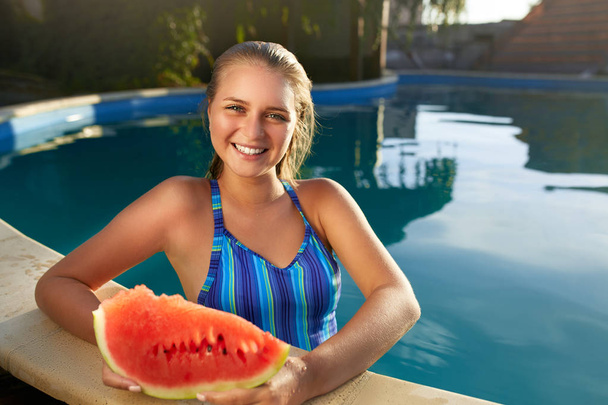 Pretty tanned slim young woman holds slice red watermelon over blue pool, relaxing on tropical island in resort, eating healthy food. Fruit summer diet. Vegetarianism and vegan lifestyle. - 写真・画像