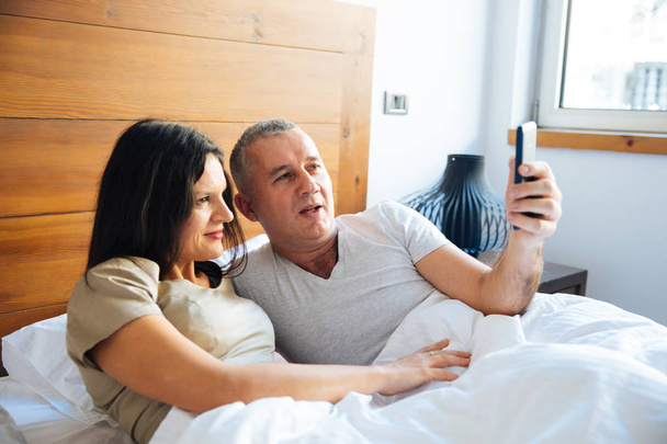 attraktives und glückliches Paar mittleren Alters, das morgens im Bett genießt, ein Smartphone benutzt und ein Selfie macht - Foto, Bild