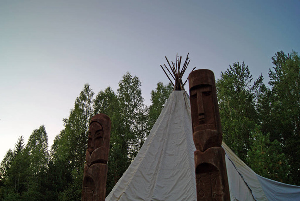 dos tótems de madera paganos frente a una carpa tradicional (chum o tipi) primer plano sobre un fondo de primer plano del bosque
 - Foto, Imagen