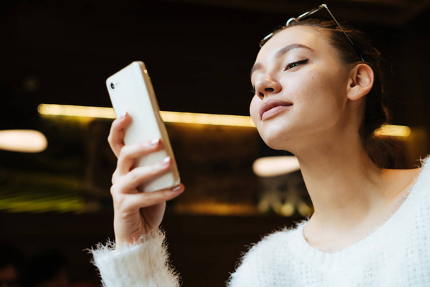 confident beautiful girl looks into her smartphone and smiles, sits in a cafe - Foto, Bild