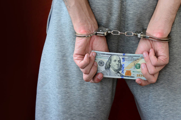 Ein Festgenommener in grauer Hose mit gefesselten Händen hält eine riesige Menge Dollarscheine in der Hand. zurück. - Foto, Bild