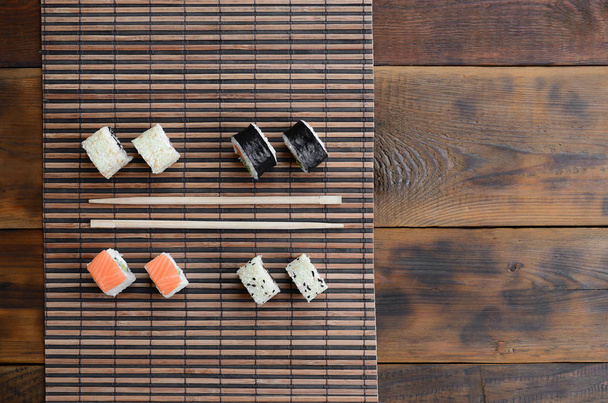 Роллы суши и деревянные палочки лежат на коврике из бамбуковой соломы. Традиционная азиатская кухня. Вид сверху
. - Фото, изображение