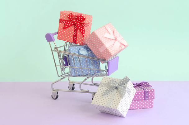 Small gift boxes of different colors with ribbons lies in shopping cart on a violet and blue pastel background. - Foto, Imagem