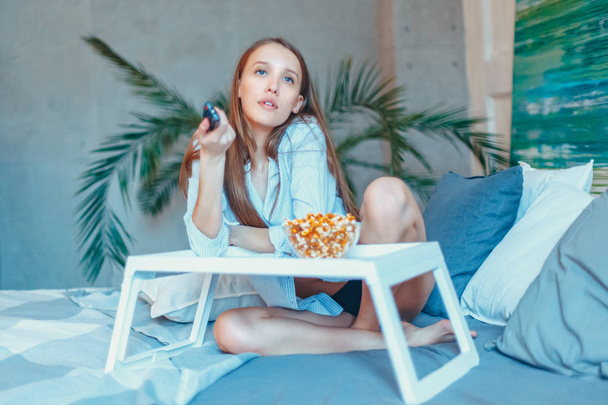 Beautiful young smiling woman watching a movie in the bed and eating popcorn. - Φωτογραφία, εικόνα