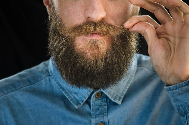 Bearded Man in a Blue Denim Shirt Twirling His Mustache with His Fingers. Portrait of a Hipster on Black Background Closeup - Fotografie, Obrázek