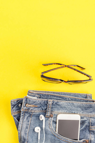Плоская накладка синих джинсов и наушников смартфона на жирном желтом бумажном фоне с копировальным пространством. Вид сверху на женщину в повседневной одежде. Trendy hipster look top view
 - Фото, изображение