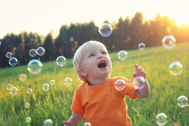 Cute toddler blond boy playing with soap bubbles on summer field. Happy childhood concept. Authentic lifestyle image. - Photo, Image