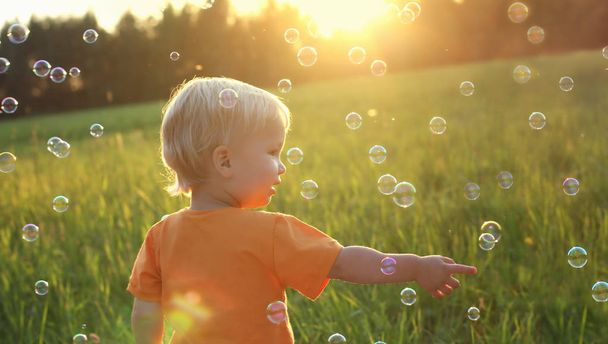 Blondýna Roztomilé batole chlapec hraje s mýdlovými bublinami na letní pole. Krásný západ slunce světlo. Pojem šťastné dětství. Autentické životní styl obrázek. - Fotografie, Obrázek