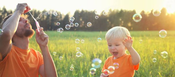Schattig peuter blonde jongen en zijn vader spelen met zeepbellen op zomer veld. Prachtige zonsondergang licht. Gelukkige jeugd concept. Authentieke levensstijl afbeelding. - Foto, afbeelding