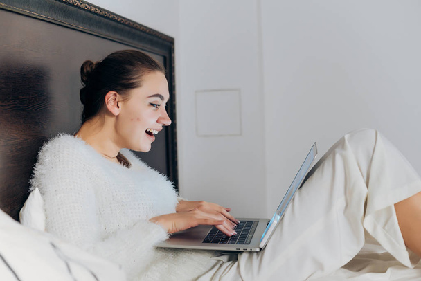 ευτυχισμένος κορίτσι γέλιο με λευκά ρούχα, κάθεται στο κρεβάτι και κοιτάζοντας αστεία σειρά για το laptop - Φωτογραφία, εικόνα