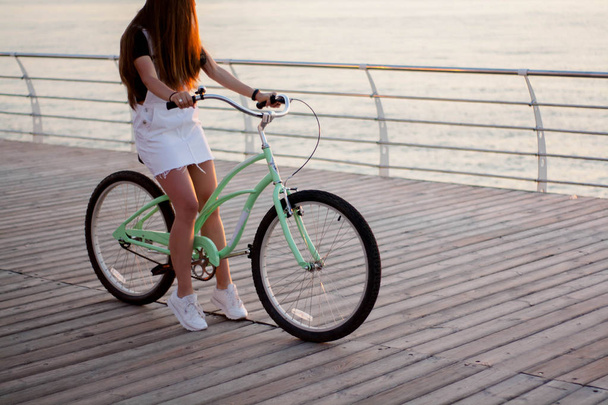 Beautiful longhaired young woman with a bicycle having fun, cycling, posing on the camera near the sea on the sunrise - Фото, зображення