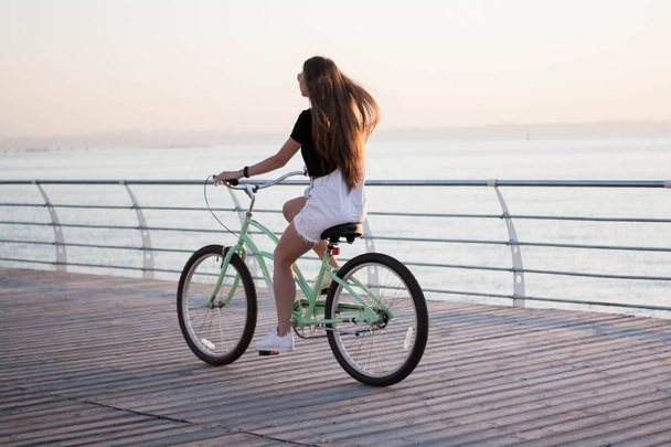 Bela jovem de cabelos compridos com uma bicicleta se divertindo, pedalando, posando na câmera perto do mar no nascer do sol
 - Foto, Imagem