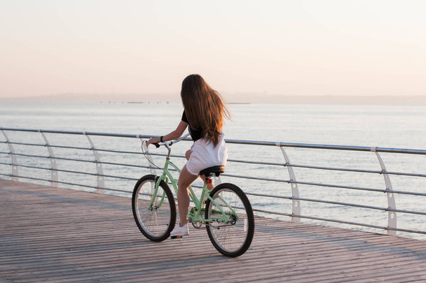 Hermosa mujer joven de pelo largo con una bicicleta que se divierte, ciclismo, posando en la cámara cerca del mar en el amanecer
 - Foto, Imagen