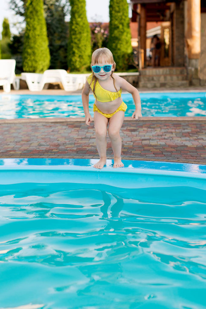 Kleines Mädchen in gelbem Badeanzug und Sonnenbrille schwimmt und springt zum Schwimmbad im Resort. Sommerspaß im Schwimmbad.  - Foto, Bild