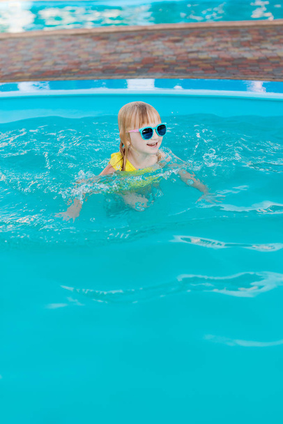 Kleines Mädchen in gelbem Badeanzug und Sonnenbrille schwimmt und springt zum Schwimmbad im Resort. Sommerspaß im Schwimmbad.  - Foto, Bild