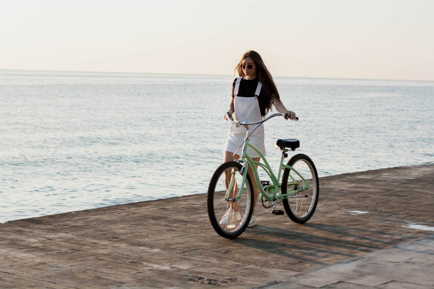 Beautiful longhaired young woman with a bicycle having fun, cycling, posing on the camera near the sea on the sunrise - Photo, Image