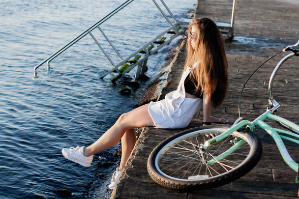 Beautiful young woman in sunglasses with a bicycle is resting near the sea, having fun, looking at a beautiful view at sunrise and posing on camera - Foto, imagen