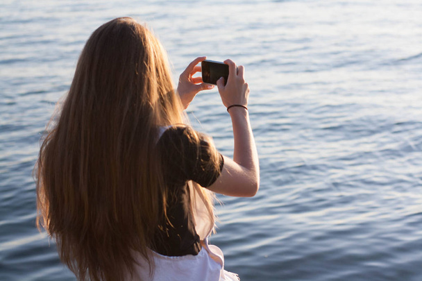 Mooie jonge vrouw met lange haren heeft leuke maakt gebruik van een smartphone, noemt, schrijft sms, surfen op internet, maakt selfy bij zonsopgang - Foto, afbeelding