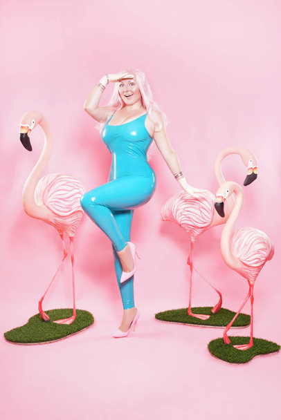 Sevimli tombul kız pembe studio arka plan üzerinde büyük bir Flamingo ile poz parlak mavi lateks mayo giymiş - Fotoğraf, Görsel