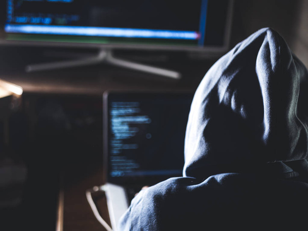hacker in hoodie typing code in program using laptop in dark studio - Photo, image