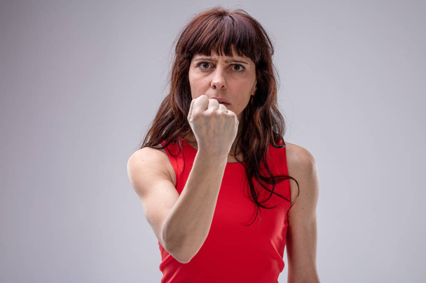 Mujer enojada haciendo un gesto con su puño cerrado a la cámara con una expresión implacable feroz sobre gris
 - Foto, Imagen