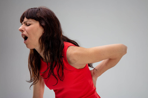 Mulher com dor nas costas gemendo de dor enquanto segura a mão na parte inferior das costas em uma visão lateral de perto no cinza
 - Foto, Imagem