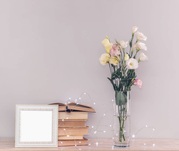 Eustoma flowers bouquet in a vase, stack of old vintage books, white photo frame and garland lights on a grey background. Reading and relax concept. - Foto, afbeelding