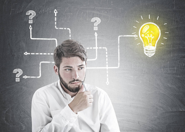 Troubled young bearded caucasian businessman wearing a white shirt sitting with pen near his face and thinking looking sideways. A bright colorful light bulb with tangled arrows drawn on a blackboard - Photo, Image