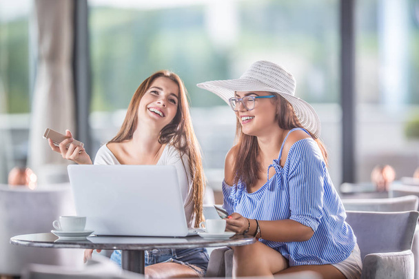 Two young cheerful women in a coffee shop with a credit card and and laptop comfortably buy them over the internet. - Foto, Imagen