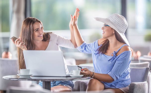 Two young cheerful women in a coffee shop with a credit card and and laptop comfortably buy them over the internet. - 写真・画像