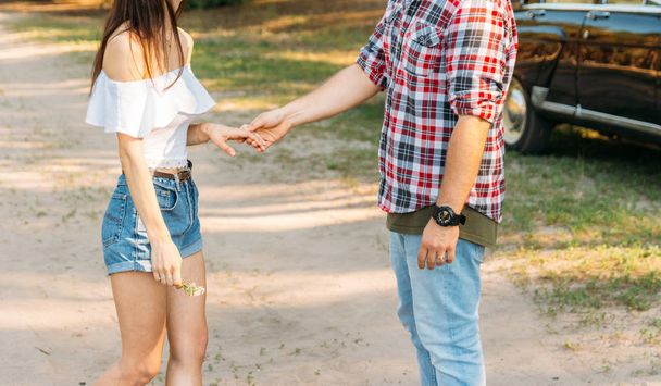 the guy is going through, modestly, in a plaid shirt and jeans, a farmer holding a girl in denim shorts and a white blouse, holding her hand, talking, making a marriage proposal. takes the girl by the hand.in the background is a black car.offering - Foto, afbeelding