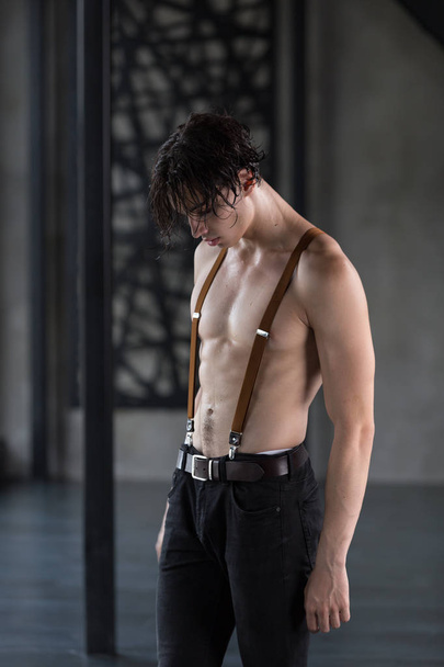 Handsome shirtless dark haired male model looking down in ripped jeans and suspenders in a dark room - Zdjęcie, obraz
