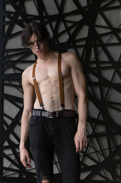 Handsome shirtless dark haired male model in ripped jeans and suspenders in a dark room - Valokuva, kuva
