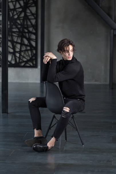 Stylish handsome young man with wet dark hair in all black outfit with turtleneck and ripped jeans sitting on a chair with crossed arms looking over a shoulder in a dark room - Foto, Bild