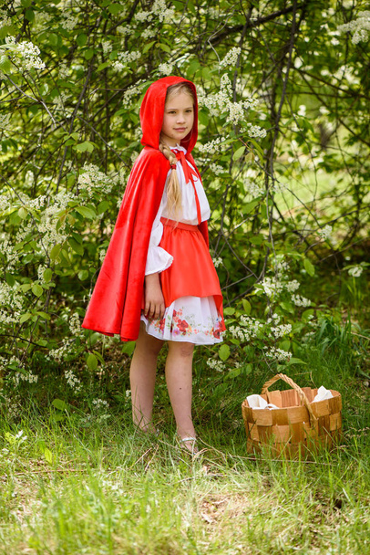 Chica rubia sonriente posando en un vestido de capucha roja cerca de árboles verdes con flores blancas
 - Foto, Imagen