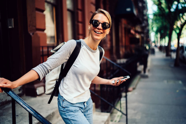 Half length portrait of cheerful young woman leaving building laughing holding telephone,smiling hipster girl in sunglasses spending weekends actively in good mood strolling in downtown with smartphon - Foto, afbeelding