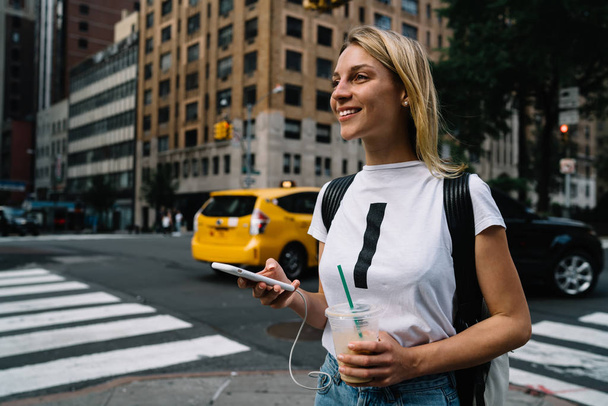 Cheerful young female tourist walking on city street passing cars and crosswalk enjoying big city,smiling hipster girl excited with fast taxi service calling cab via application on smartphon - Foto, afbeelding