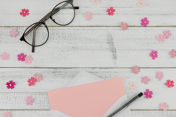 leere rosa Karte in weißem Umschlag, Stift und Brille dekorieren mit rosa Papierblumen auf weißem Holzhintergrund mit Kopierraum - Foto, Bild
