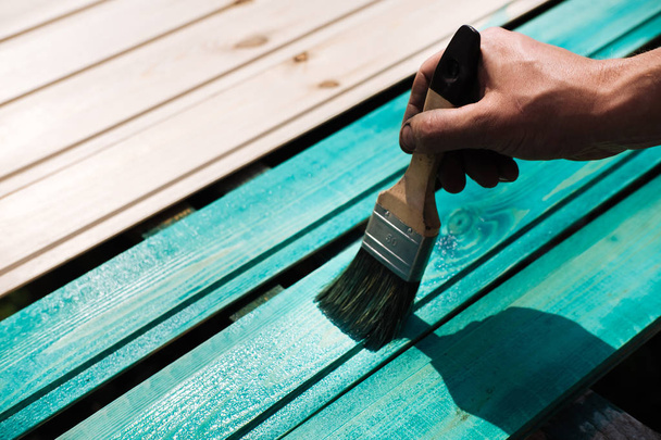 Coating with colored protective paint on the wooden surface of the lining boards. Hand with a brush paints a bright turquoise surface outdoors in Sunny weather. - Photo, Image