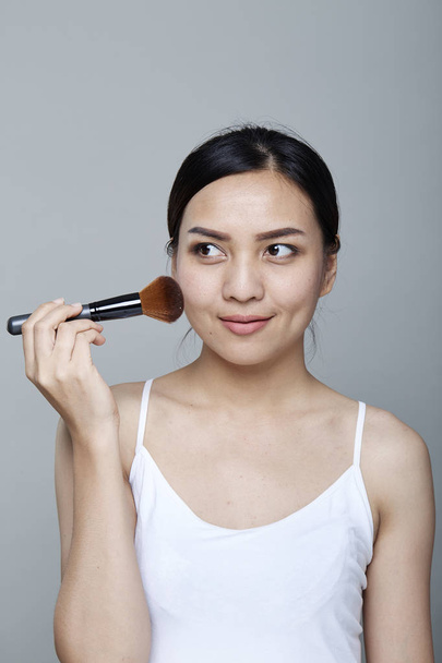 Piękna dziewczyna azjatyckich stosowania makijażu na szarym tle, Portret młodej kobiety w beauty salon wnętrza. Stosując odcień skóry. - Zdjęcie, obraz