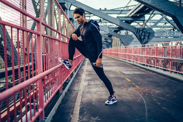 Young man in active wear listening music in earphones and warming up before training on modern bridge.Sportive man in black tracksuit and sport sneakers having workout outdoors while enjoying songs - Φωτογραφία, εικόνα