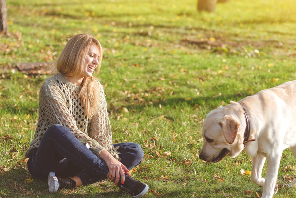 beautiful blonde is sitting on the grass in the park and playing with her dog labrador - Zdjęcie, obraz