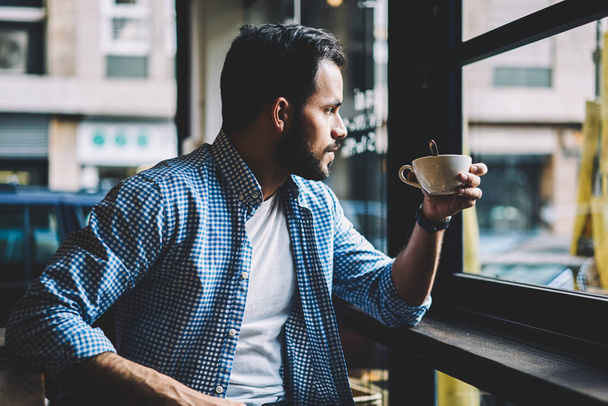 Pensive bearded hipster guy enjoying morning coffee for breakfast looking away through window in cafeteria, thoughtful caucasian male in casual outfit drinking beverage during break in cafe interior - Photo, image