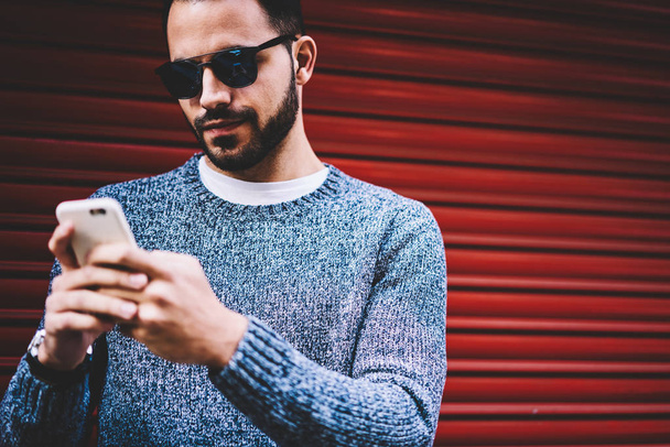 Handsome young man sending text message chatting on smartphone standing on urban settings background,hipster guy checking notification on cellular updating application spending free time outdoors - Фото, изображение