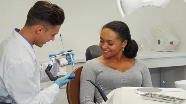 Professionele tandarts iets aan zijn vrouwelijke patiënt terwijl kunstgebit uit te leggen. Mooie Afrikaanse vrouw en haar tandarts bespreken tandheelkundige schimmel. Gezondheid, geneeskunde, dienstverleningsconcept. - Video