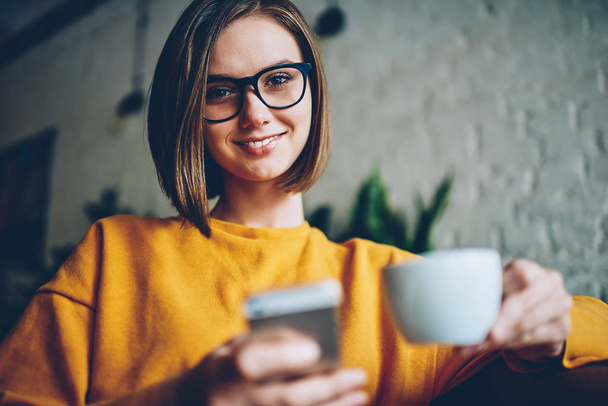 Half length portrait of attractive successful female entrepreneur in stylish eyeglasses with black frame smiling at camera resting in cozy coworking space with cup of tasty coffee and digital smartphone - Zdjęcie, obraz
