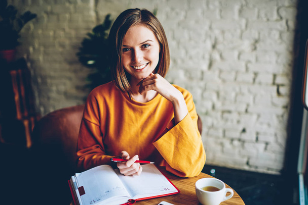 Half length portrait of beautiful cheerful student with stylish short haircut smiling at camera making notes in notebook at cup of coffee in morning time.Positive female owner writing working plan - Zdjęcie, obraz