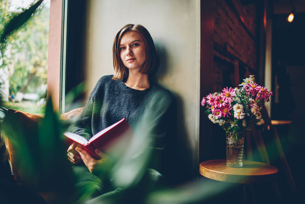 Half length portrait of attractive female reader with short haircut looking at camera spending recreation time on favourite book.Charming smart student resting in coworking and enjoying bestseller - Zdjęcie, obraz